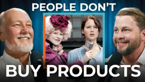 Three people hold microphones in front of a "Hunger Games" scene with the text: "People Don't Buy Products," discussing buying psychology and consumer identity's impact on sales.